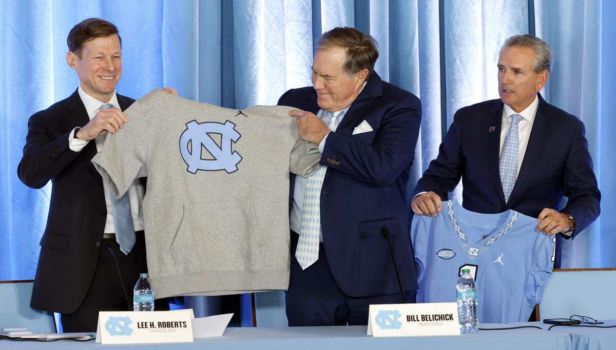 UNC Chancellor Lee Roberts gives new North Carolina head football coach Bill Belichick a hoodie with cut off sleeves during a press conference at the Loudermilk Center at UNC in Chapel Hill, N.C., Thursday, Dec. 12, 2024. UNC AD Bubba Cunningham stands to the right.
