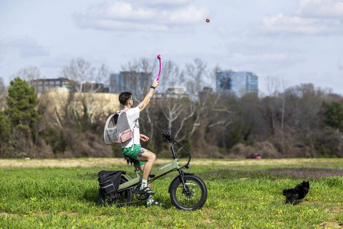 Andrew Gregg plays fetch with his 6-year-old rescue dog, Bubs, while riding his e-bike at Dorothea Dix Park with the Raleigh skyline in the background on Thursday, March 5, 2026, as temperatures climbed into the upper 70s during one of the final days of winter. Gregg says Bubs often rides in a pet carrier backpack with him on outdoor adventures when the weather is nice.