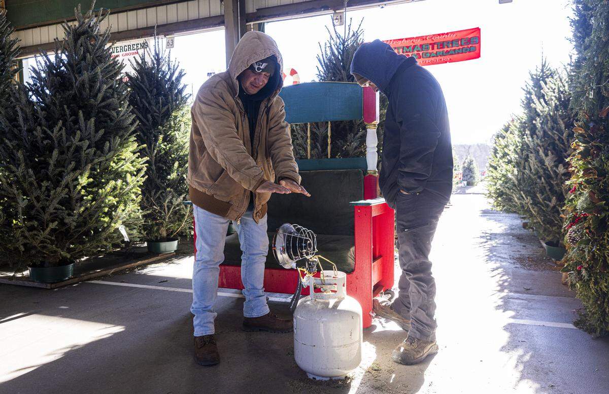 Brothers George, left, and Israel Delgado of Jernigan’s Nursery in West Jefferson try to keep warm near a propane heater at the company’s Christmas tree vendor stall at the North Carolina State Farmers Market in Raleigh on Monday morning, Dec. 15, 2025. Temperatures in Raleigh dipped into the teens, marking the city’s coldest weather since the winter of 2022.