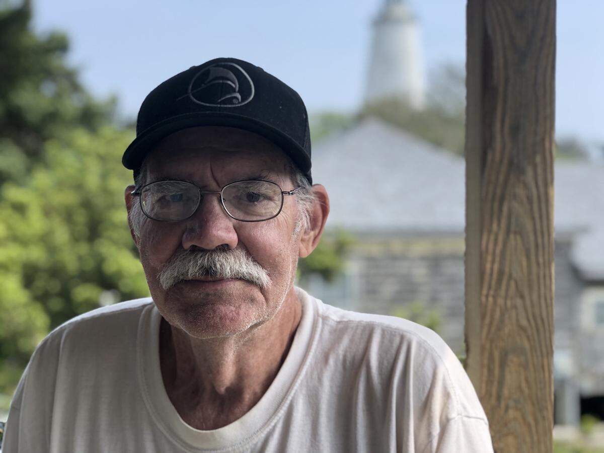 Clayton Gaskill on the porch the rental house he’s rebuilding after it was flooded by Hurricane Dorian in 2019. Behind him are the house where he lives, and the Ocracoke Light.