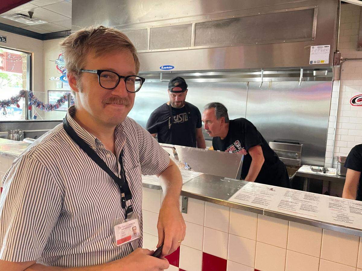 News & Observer food writer Drew Jackson at Cloos’ Coney Island in the Mission Valley Shopping Center near NC State’s campus in Raleigh, July 11, 2024. Owner Daniel Cloos is shown working in the background, on the right.