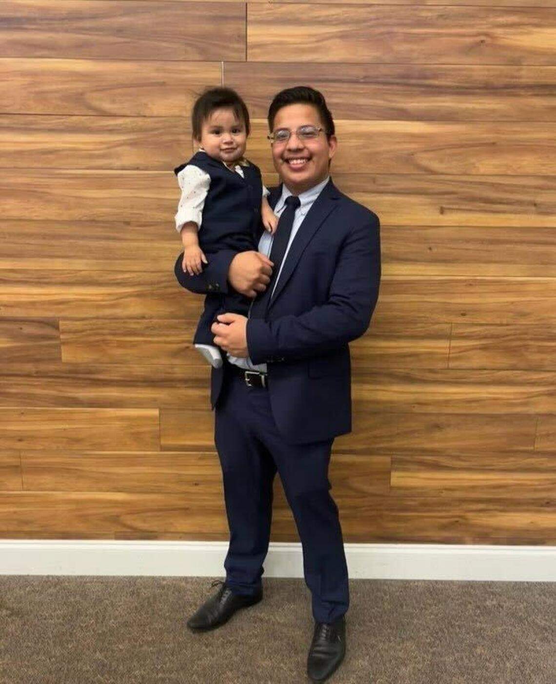 Moises Benitez Diaz poses for a portrait photo behind a brown wooden wall while holding his son, Elijah. Elijah is wearing a dark blue vest over a white shirt while Benitez Diaz wears a navy suit and tie.