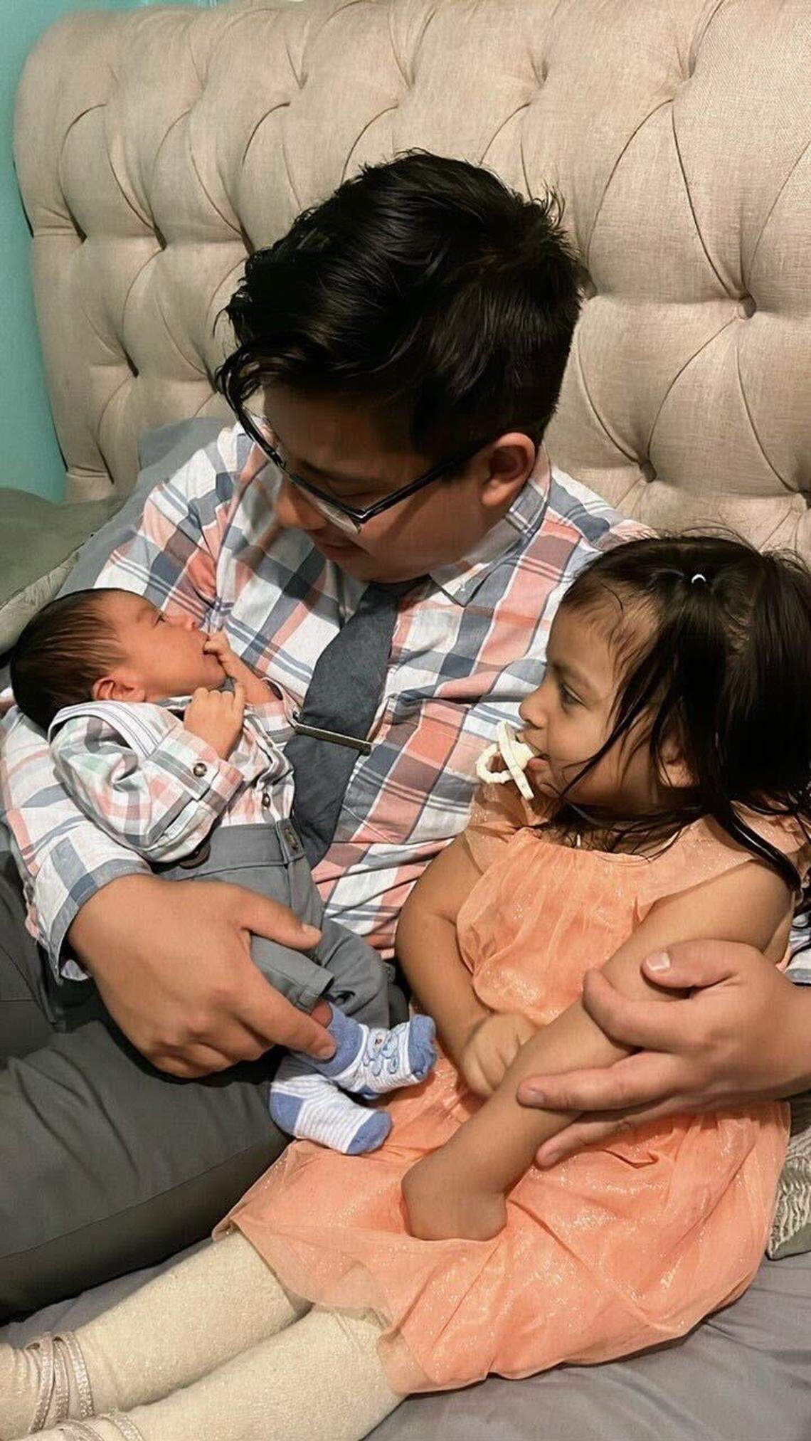 Moises Benitez Diaz sits on a white leather couch with his two kids, Rochelle and Elijah. He and Elijah are wearing matching white plaid button-up shirts and gray dress paints, while Rochelle wears an orange dress.