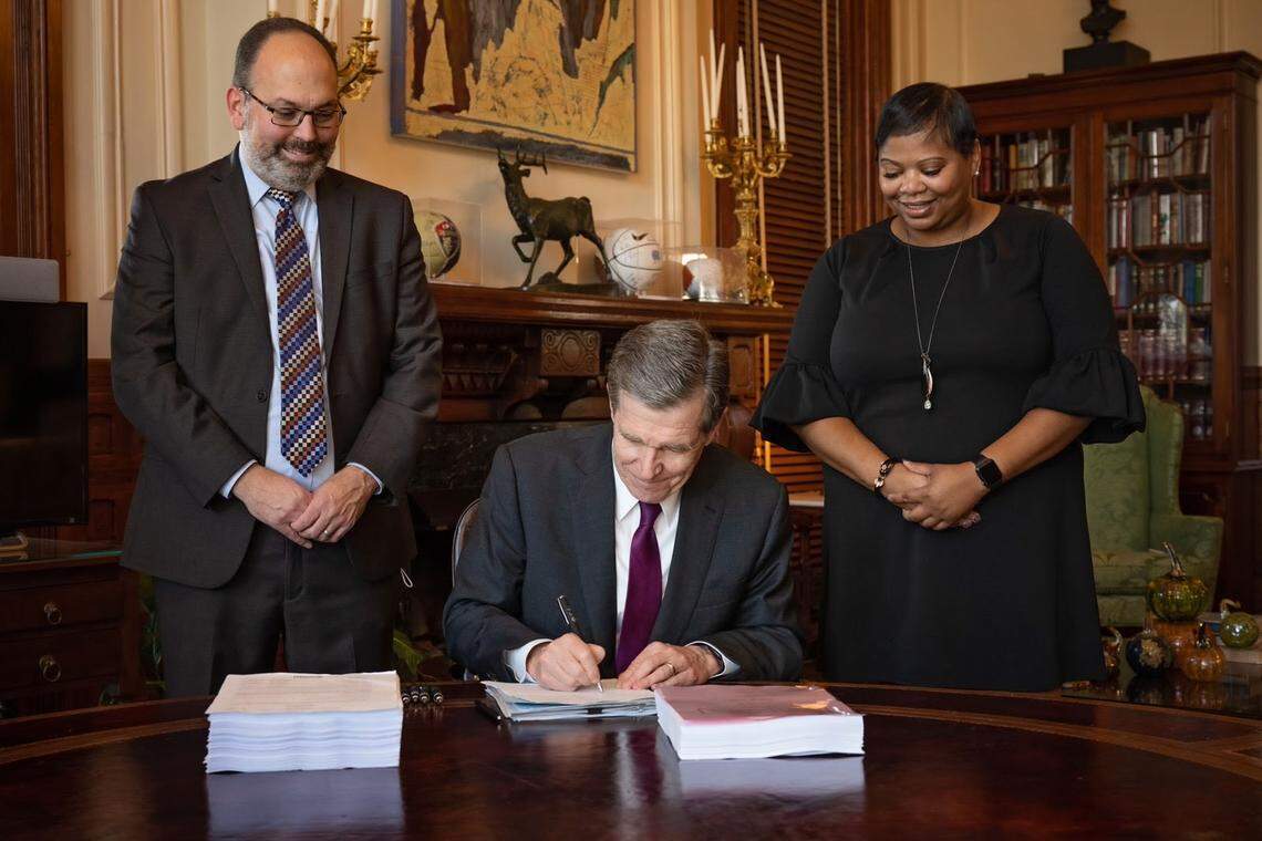 North Carolina Gov. Roy Cooper signs the state budget into law on Thursday, Nov. 18, 2021, flanked by state budget director Charlie Perusse and L.T. McCrimmon, who is director of Legislative Affairs.