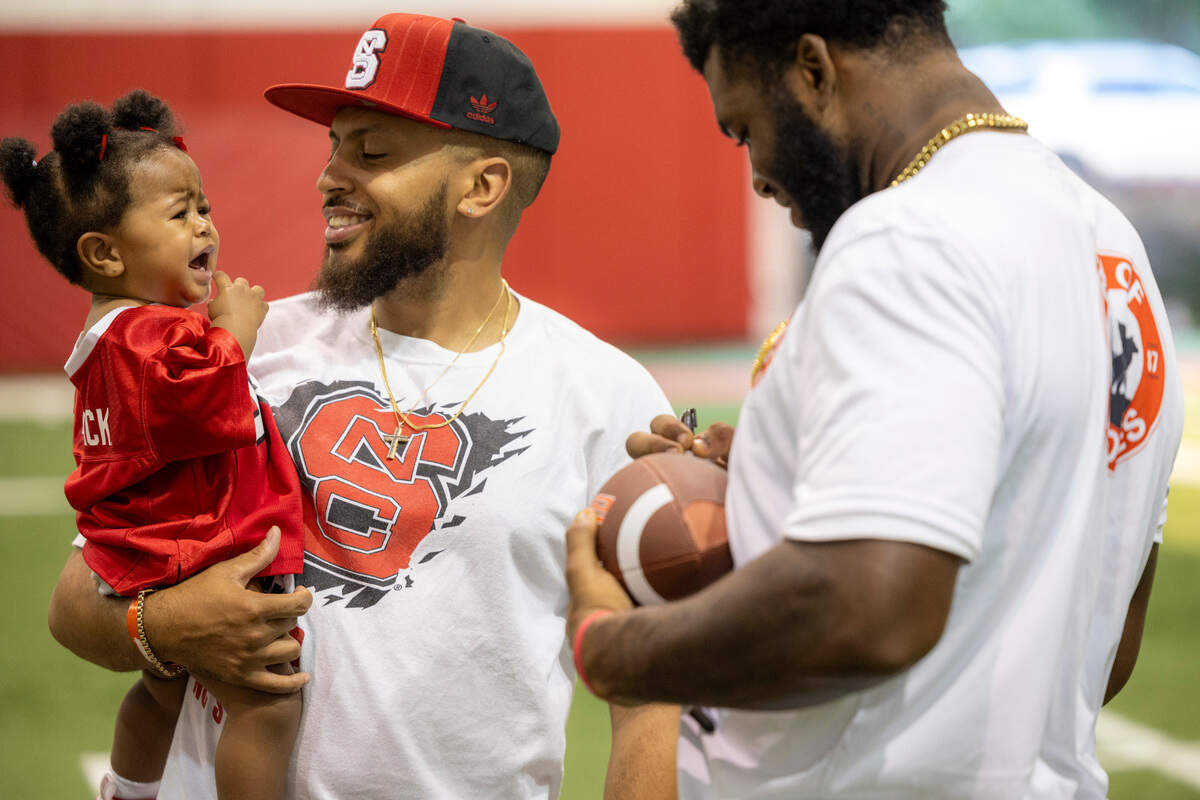 Jacoby Pulley holds his daughter Ivy Pulley, 1, as offensive lineman Jacarrius Peak autographs a football during N.C. State’s ‘Meet the Pack’ event with players, fans at the Close-King Indoor Practice Facility Saturday, Aug. 16, 2023.