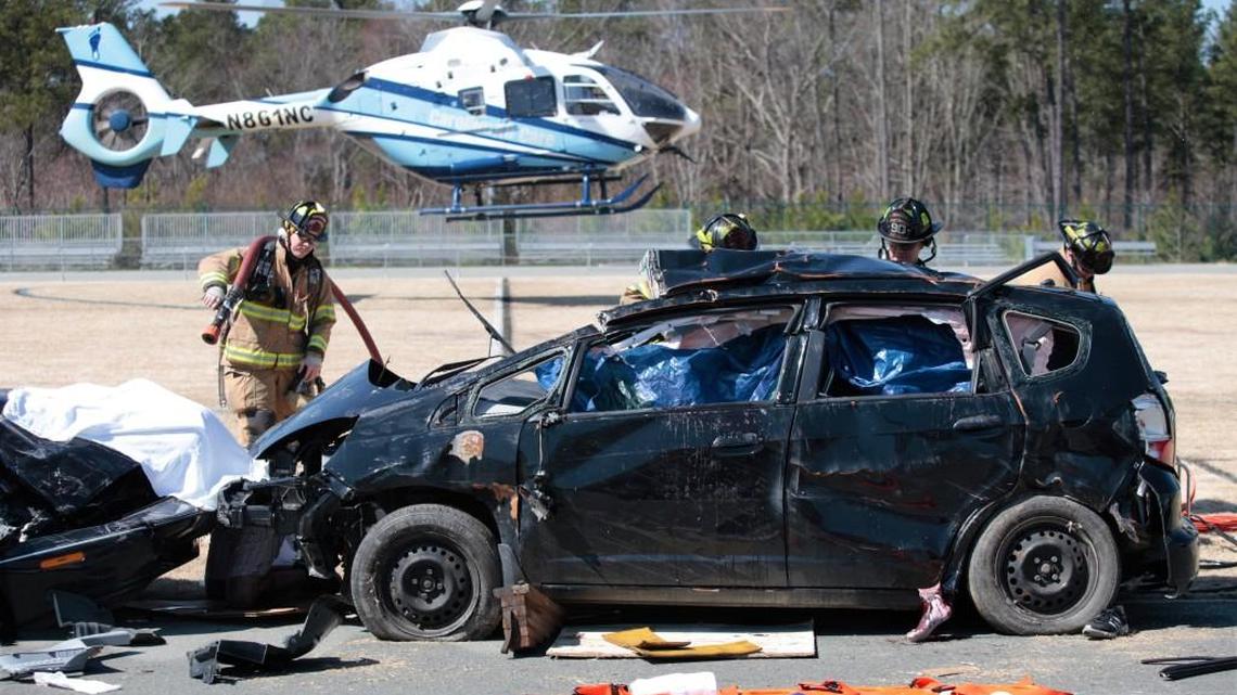 For realism in a mock car crash for Carrboro High School students Friday, March 21, 2014, a Carrboro firefighter, left, stands by with a water hose as fellow Carrboro firefighters work to extricate Carrboro High student volunteers acting the part of car crash victims on the CHS football field. The Tarheel One rescue helicopter and crew lift off in background with a simulated patient. Over 500 CHS juniors and seniors watched as real local first responders attend victims of a mock head-on collision to illustrate the dangers of impaired and distracted driving.