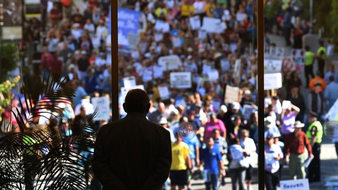 A member of the NC General Assembly sergeant at arms watches as Moral Monday protesters cross the street towards the Legislative Building on May 16, 2016. Hundreds of protesters gathered inside and outside the N.C. Legislative Building to protest the controversial House Bill 2.