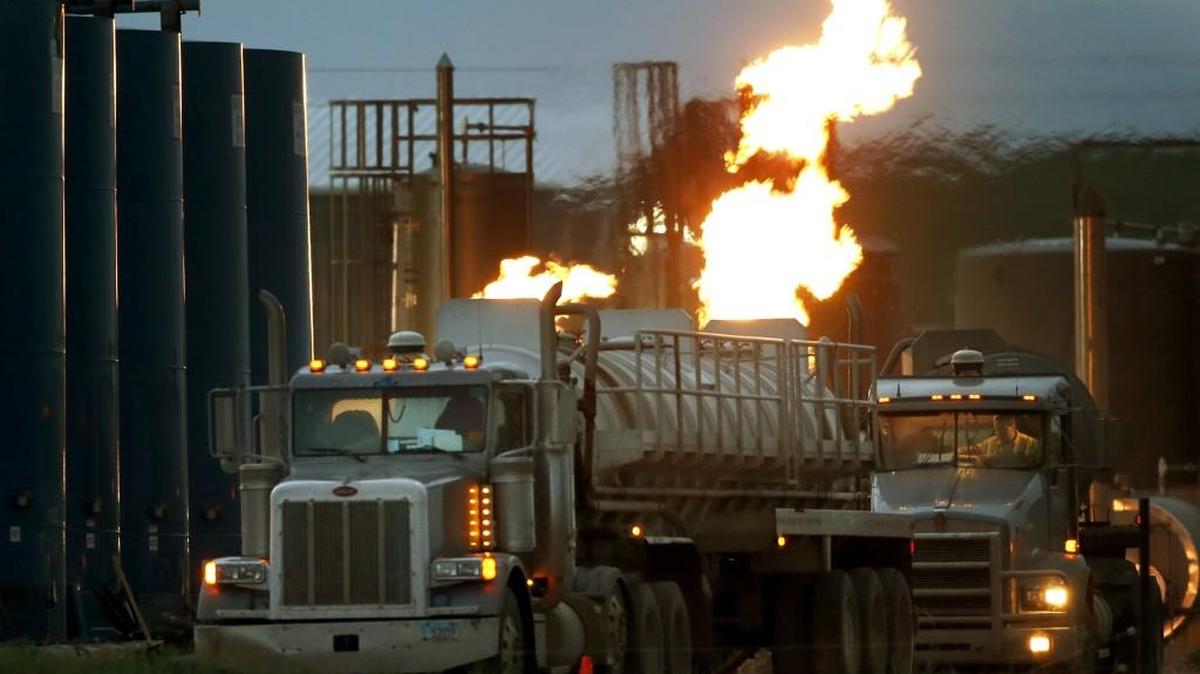 In this June 9, 2014 photo, drivers and their tanker trucks, capable of hauling water and hydraulic fracturing liquid, line up near a natural gas burn off flame and storage tanks in Williston, N.D.