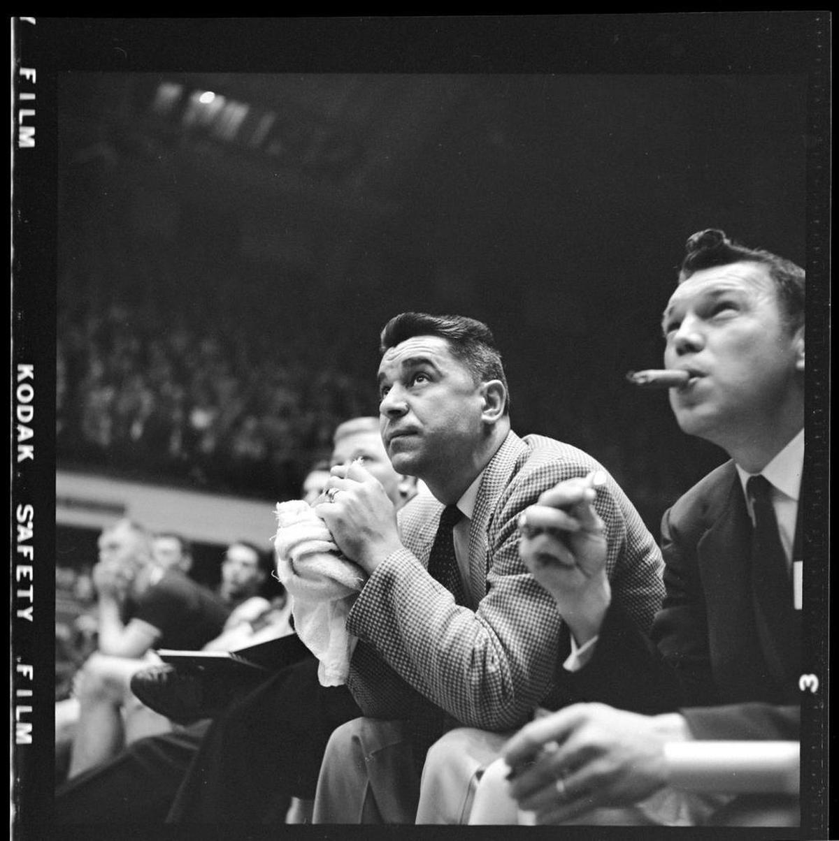 An assistant coach lights up a cigar on the bench (right) as Clemson coach Press Maravich works his trademark towel during a game in the 1957 ACC Tournament in Reynolds Coliseum.